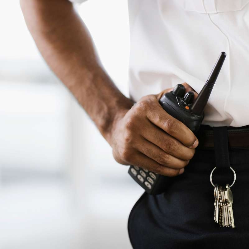 A person in a white shirt and dark pants, representing Private Security Oakland County, holds a walkie-talkie with keys dangling from their belt. The background is artfully blurred, highlighting the communication device and keys, essential tools for maintaining safety in Oakland County events.