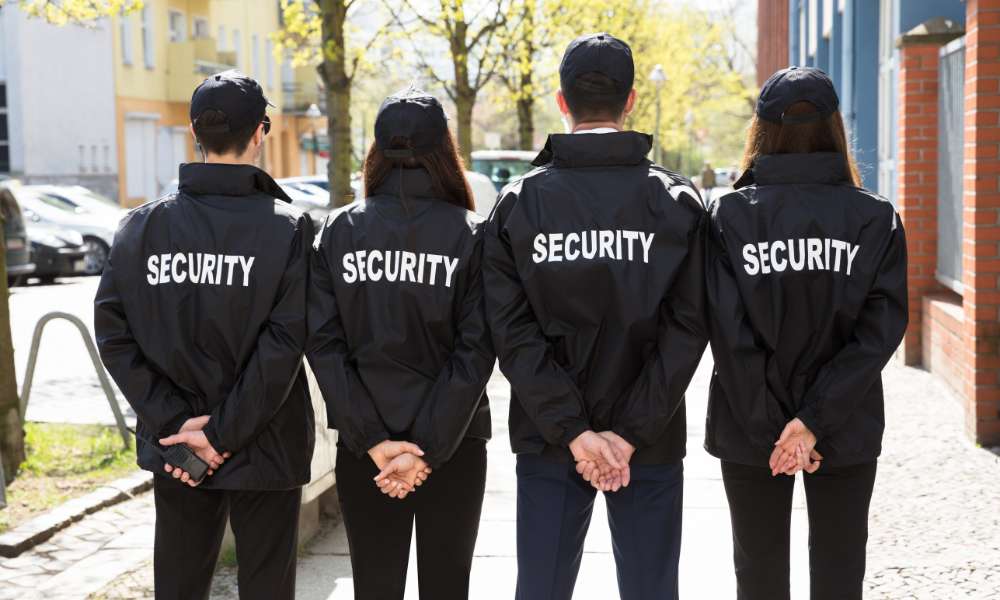 On a sunny tree-lined sidewalk, four security guards in black jackets and caps, emblazoned with "SECURITY," stand with hands behind their backs. Representing Private Security Oakland County, they maintain a vigilant presence to ensure safety and order.