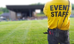 A person in a bright yellow "Event Staff" shirt, representing Special Event Security Oakland County, stands with their back to the camera, overseeing a large crowd at an outdoor event. Equipped with a radio and tools on their belt, the scene takes place on a grassy area.