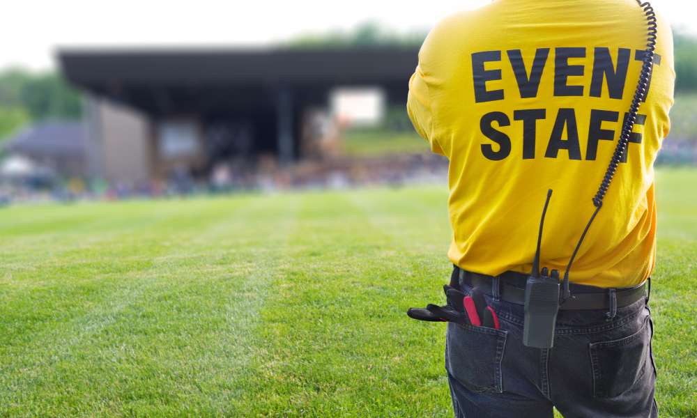 A person in a yellow "Event Staff" shirt stands on a grassy field with a two-way radio and tools clipped to their belt, overseeing an events stage in the distance.