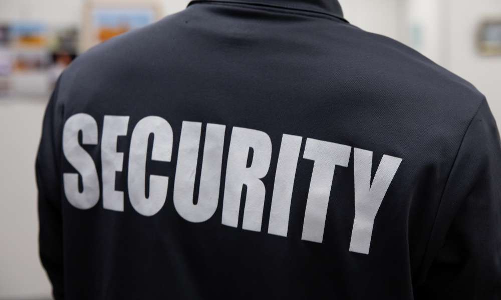 Back view of a person wearing a black jacket with the word "SECURITY" printed in large white letters, standing near places of worship.