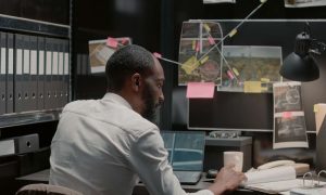 A man sits at a cluttered desk in a dimly lit room, looking at his laptop. The wall, reminiscent of an investigation board, features photos and notes linked by strings. Amid scattered office supplies, a lamp casts its glow on this private security consultant from Oakland County deep in thought.