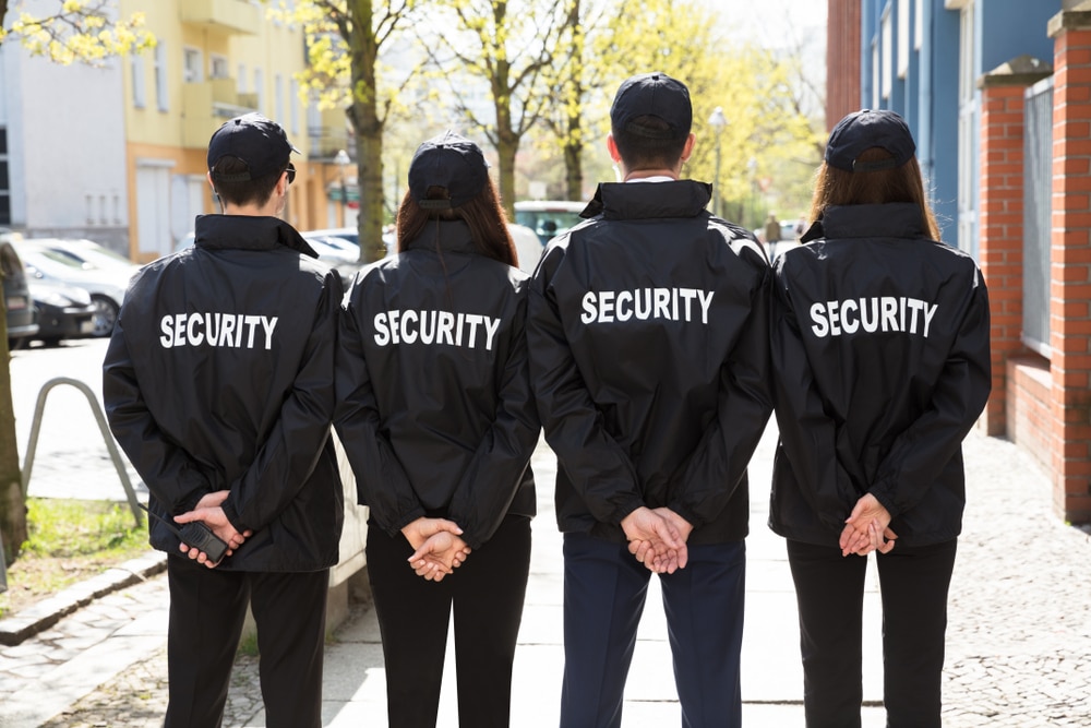 Four security guards in black jackets and caps, with "SECURITY" printed on their backs, stand in a line on a city sidewalk. Representing top-tier Security Services Oakland County, they have their hands behind their backs, facing away from the camera. A street with buildings and trees is visible.