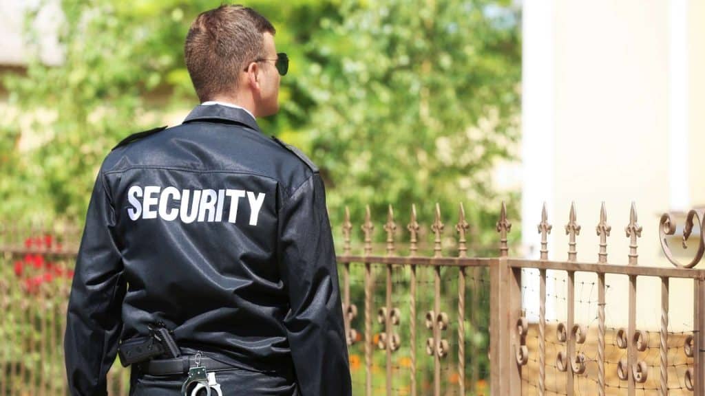 A security guard in a black uniform and sunglasses stands in front of a wrought iron fence, facing away from the camera with greenery in the background.