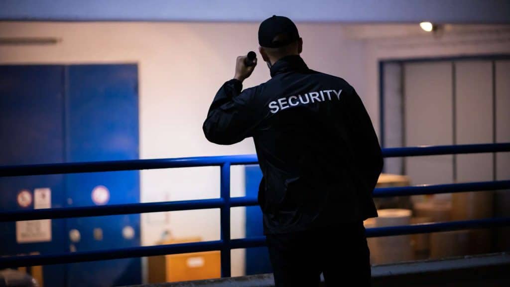 A security guard in a black jacket and cap, with "SECURITY" on the back, holds a flashlight. The scene is dimly lit, featuring blue doors and a railing in the foreground—a typical setting for Special Event Security in Oakland County ensuring safety and peace of mind.