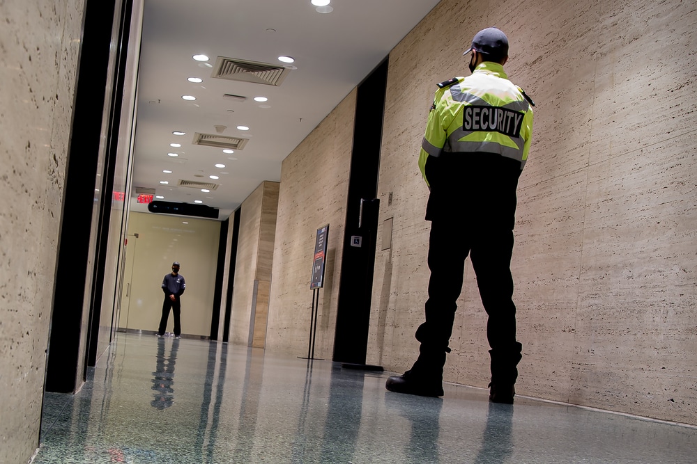 A security guard in a bright uniform stands in a well-lit hallway with marble walls and a polished floor, courtesy of a top Security Company in Oakland County. Another person is seen in the distance near a doorway, with various signs visible along the corridor.