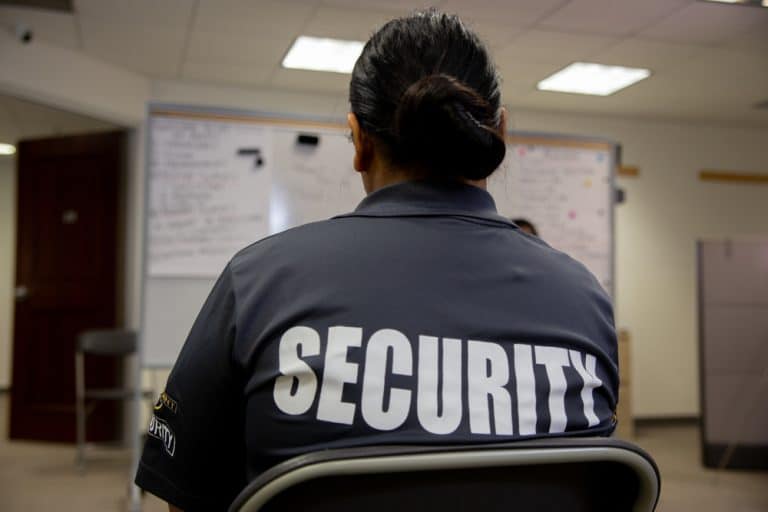 A person with dark hair in a bun is seated, wearing a gray "SECURITY" shirt. They are part of the Special Event Security Oakland County team, stationed in an office room with whiteboards covered in blurred notes behind them.