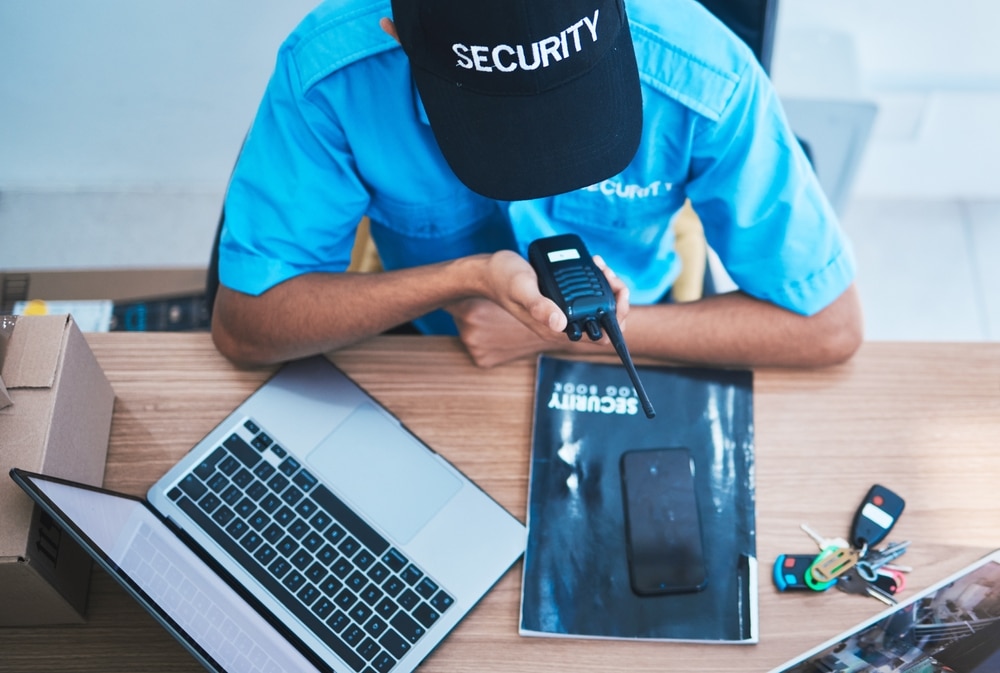 A person wearing a "SECURITY" hat and uniform uses a walkie-talkie at a desk with a laptop, keys, magazine, and a phone on it.