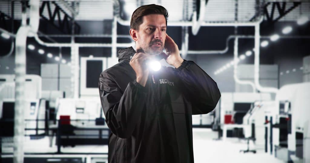 A security guard in a dark uniform holds a flashlight to his face while speaking into a walkie-talkie, standing in a brightly lit warehouse.