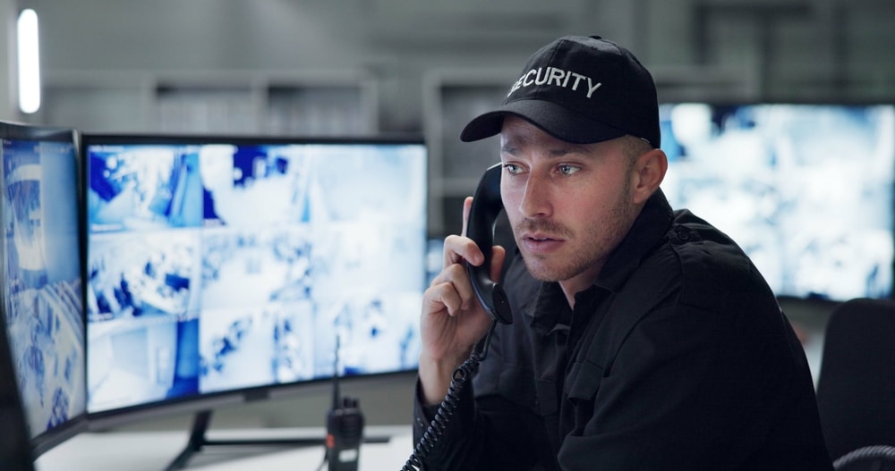 A security guard wearing a cap labeled "SECURITY" sits at a desk in an Oakland County office, holding a phone to his ear. Behind him, multiple monitors display surveillance footage, showcasing the diligence of the Private Security service engaged for monitoring operations.