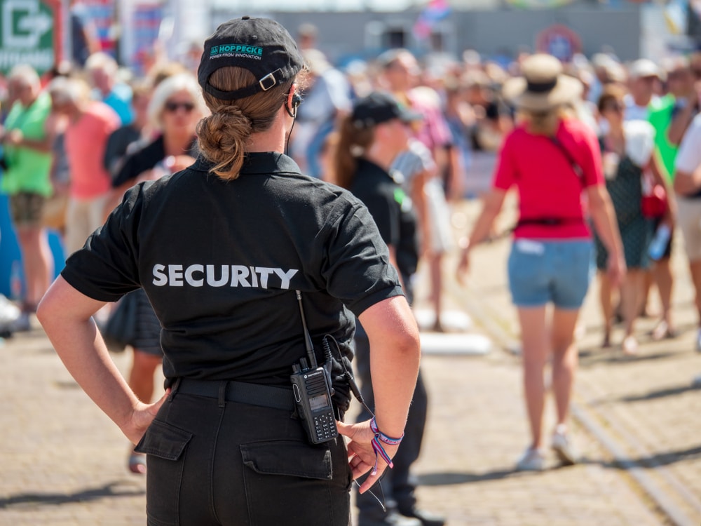 A security guard in a black uniform monitors a crowded outdoor event, with people walking around and a clear blue sky in the background.