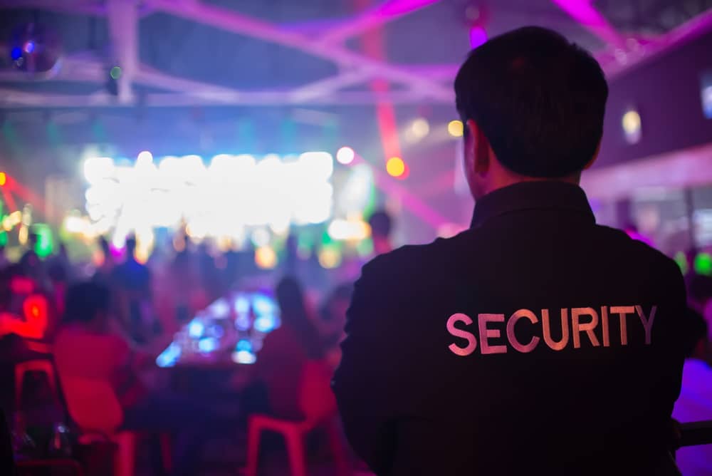 A security guard, with "SECURITY" on the back of their shirt, watches over a crowd in a brightly lit, colorful nightclub.