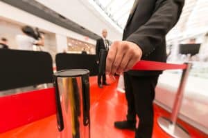 A person in a suit, part of the Event Security in Oakland County, is holding a red rope barrier on a shiny red floor. The modern interior reveals another suited individual alongside a camera on a tripod.