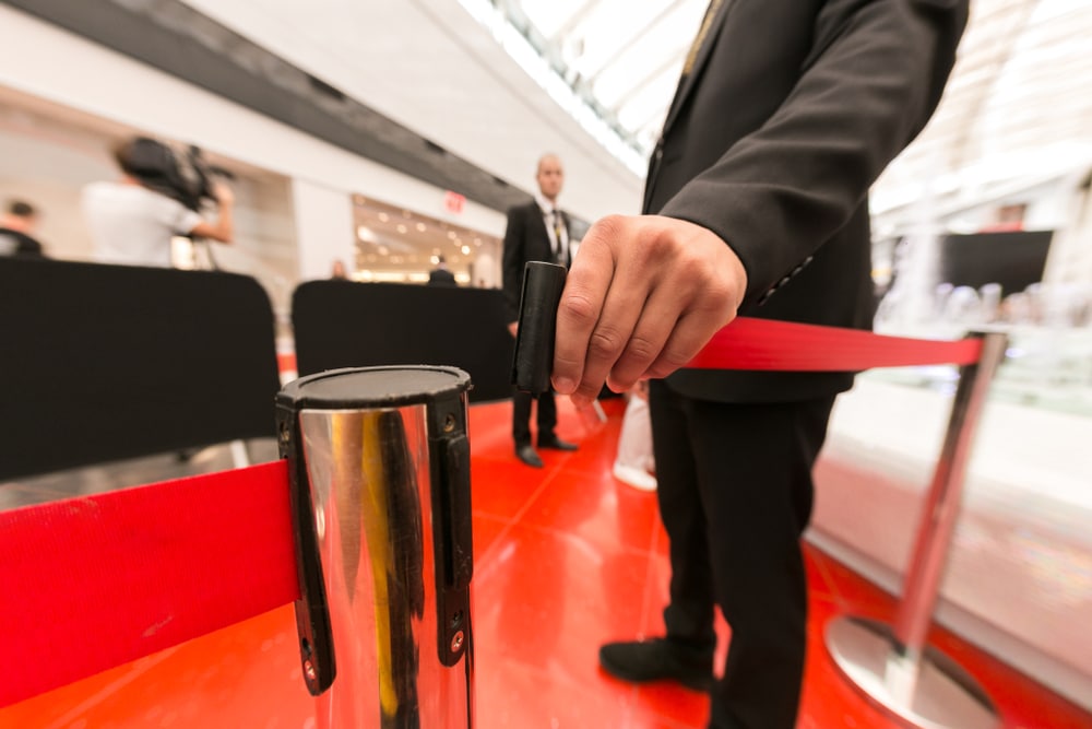 A suited person is closing a retractable belt barrier in a bright, modern indoor setting. Another person stands blurred in the background.