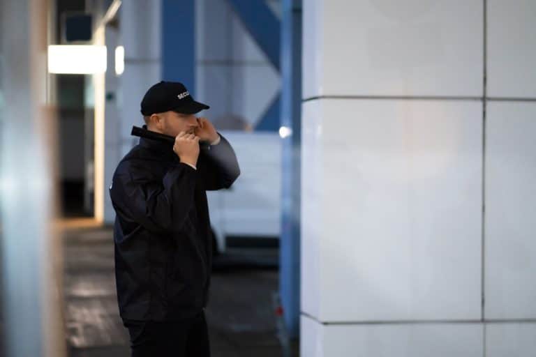 Person in black jacket and cap standing indoors, adjusting their collar. The background is slightly blurred with a white wall and some signage.