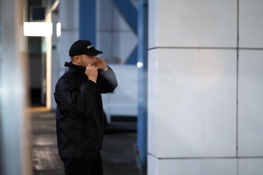 Person in black jacket and cap standing indoors, adjusting their collar. The background is slightly blurred with a white wall and some signage.