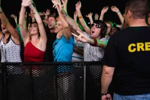 A lively crowd at a concert in Oakland County cheers in excitement near the stage barrier. People have their hands raised, some making peace signs. A person in a black "CREW" shirt, likely part of the event security team, stands in the foreground, facing the crowd.