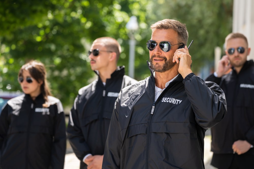 A group of four security personnel from a leading Security Company in Oakland County stand outdoors, clad in sunglasses and black jackets. The man in the foreground is speaking into a walkie-talkie. Lush greenery surrounds this scene of professional vigilance.