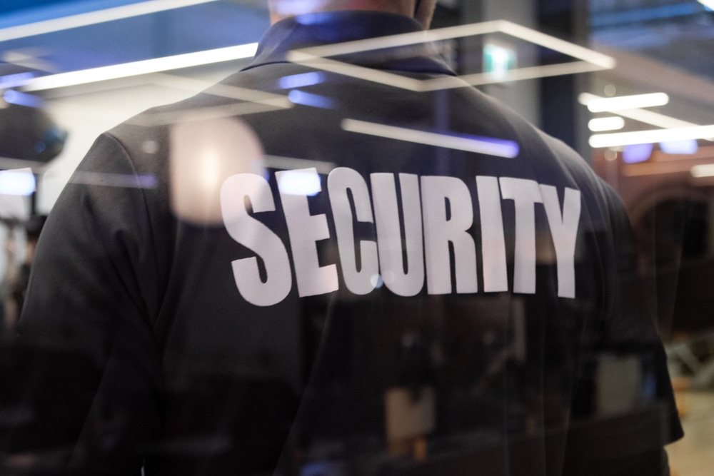 A person wearing a black shirt with "SECURITY" written in white on the back is standing in an indoor setting, symbolizing Special Event Security Oakland County, surrounded by glass reflections and blurred lights.