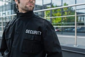 A man in a black security jacket stands outside in Oakland County, ensuring safety at a special event. The word "SECURITY" is visible on his jacket against the backdrop of a modern building with large windows and metal railings.