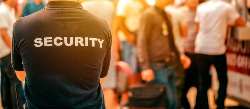 A security guard, identifiable by "SECURITY" emblazoned on their shirt, stands amidst a bustling crowd. The sunny scene showcases the vibrant activity synonymous with private security in Oakland County, highlighting its essential role in ensuring public safety and order.