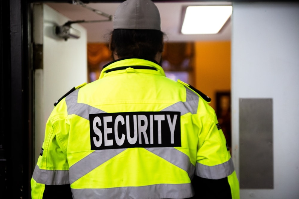 A person wearing a bright yellow jacket with "SECURITY" written on the back, standing in front of an open door.