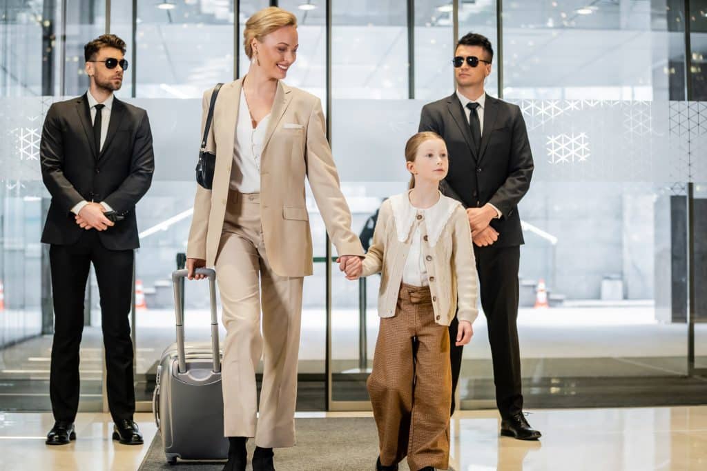 A woman in a beige suit walks hand in hand with a child pulling a suitcase, while two men in black suits and sunglasses from Security Services Oakland County stand at attention behind them in the modern building entrance.