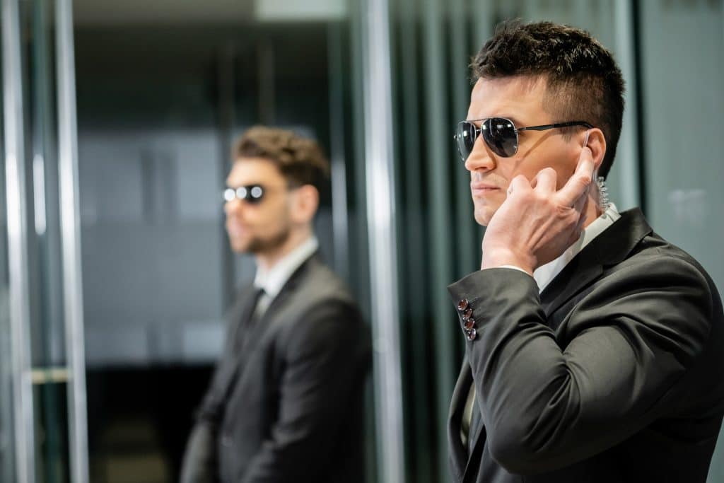 Two men in suits and sunglasses, one in the foreground touching his earpiece, stand in a professional stance, suggesting they are security personnel.