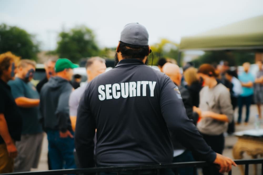 A man in a jacket labeled "Security" stands facing a lively crowd at an outdoor event in Oakland County. Clad in black with a cap, he surveys the scene where attendees socialize under a cloudy sky, surrounded by trees and tents. His presence ensures special event security for all.