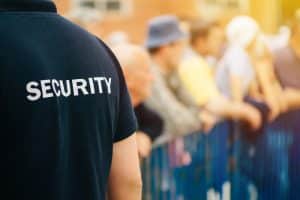 A security personnel stands in front of a crowd behind blue barriers, ensuring safety at an outdoor event with a sunny backdrop.