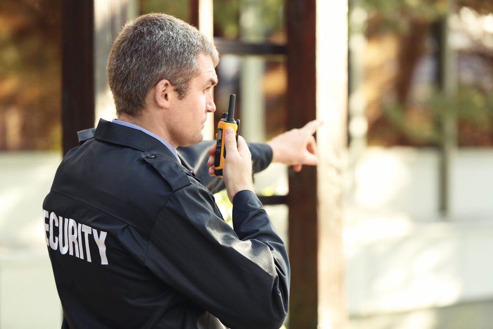 A private security guard with short hair, representing a security company in Oakland County, wears a dark uniform with "SECURITY" on the back. He holds a walkie-talkie to his mouth while pointing with his other hand, standing responsibly in a garden or park area.